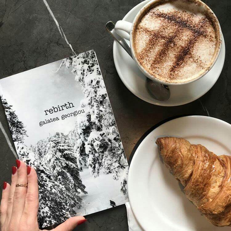 A female hand holding a copy of rebirth next to a croissant and a coffee