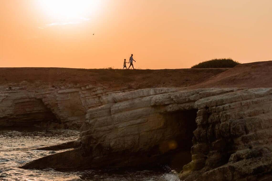 A man and a boy walk at cliffs next to the sea during sunset.